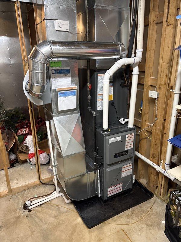 Completed full HVAC system installation including new furnace unit with integrated whole-house humidifier in residential basement utility room. Installed all necessary ductwork connections with metallic supply plenum and elbow transitions, along with white PVC condensate drainage and venting pipes properly secured to wall framing. System positioned on protective mat over concrete floor with all components mounted, connected, and ready for operation per manufacturer specifications.