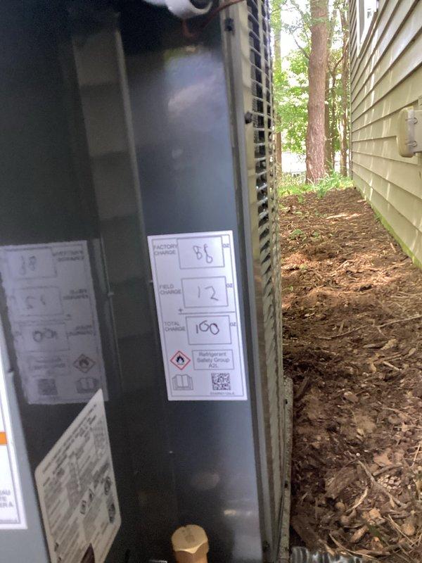 Complete residential air conditioning system replacement performed in side yard location. Removed existing outdoor condensing unit and installed new gray HVAC condenser with appropriate refrigerant line connections, including copper piping with service valves and protective coverings. Unit positioned on mulch bed against building exterior with proper clearances maintained. All manufacturer labels, warning decals, and specification plates affixed per code requirements. Refrigerant lines properly insulated and secured with flexible conduit protection. System tested and verified operational upon completion.