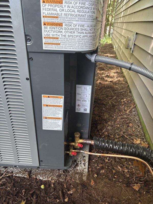 Complete residential air conditioning system replacement performed in side yard location. Removed existing outdoor condensing unit and installed new gray HVAC condenser with appropriate refrigerant line connections, including copper piping with service valves and protective coverings. Unit positioned on mulch bed against building exterior with proper clearances maintained. All manufacturer labels, warning decals, and specification plates affixed per code requirements. Refrigerant lines properly insulated and secured with flexible conduit protection. System tested and verified operational upon completion.