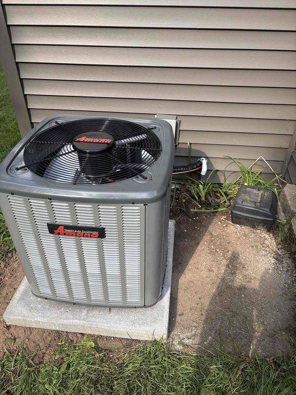 Completed full air conditioning system replacement with installation of new Amana condensing unit on existing concrete pad adjacent to building exterior. Unit properly positioned with adequate clearance for airflow and service access, connected to electrical disconnect box, and refrigerant lines terminated with brass service valves featuring protective caps. System charged per manufacturer specifications as indicated on unit nameplate, with all connections properly sealed and tested for leaks.