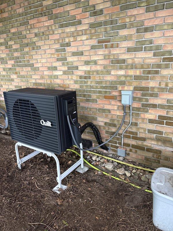 Completed installation of ductless mini-split system including outdoor condenser unit mounted on elevated stand against exterior brick wall and indoor wall-mounted air handler in interior hallway. Refrigerant lines were properly insulated and routed from outdoor unit through wall penetration, with electrical conduit and disconnect installed per code requirements.