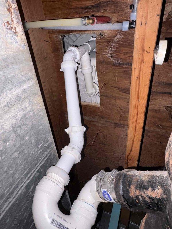 Responded to basement plumbing leak caused by drainage blockage. Inspected PVC pipe configuration beneath wooden structure and under-sink trap assembly, identifying compromised drainage system with multiple elbow joints and fittings. Cleared blockage from drain line, verified proper flow through U-trap configuration, and confirmed all connections were secure with no active leaks. Documented existing plumbing layout including PVC drainage pipes, copper fittings, and cast iron components for service records.