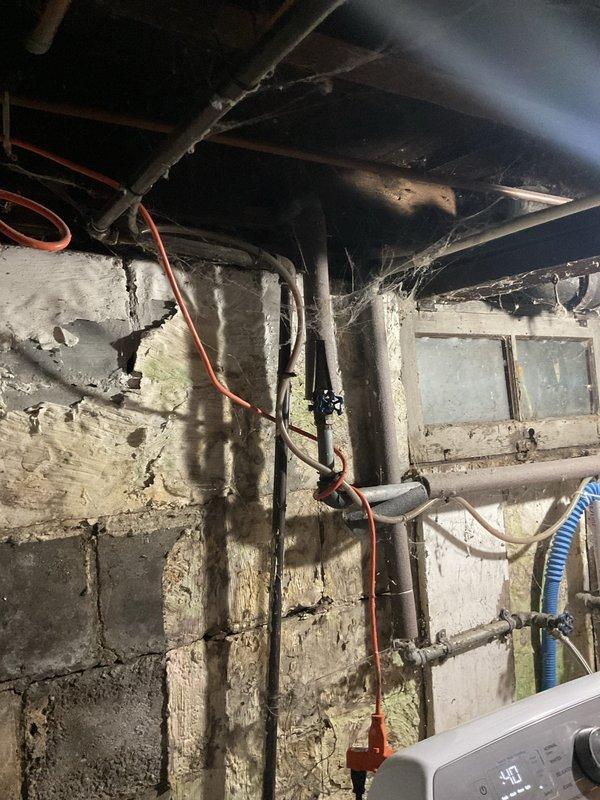 Responded to a plumbing leak complaint involving an exterior hose bib and its associated supply valve in the basement. Upon inspection, identified active leaking at the outdoor spigot mounted on the home's exterior siding, with visible water staining present below the fixture. Traced the water supply line to the basement utility area where the connecting shut-off valve was also found to be leaking. The basement environment showed evidence of long-term moisture exposure with deteriorated concrete block walls, corroded plumbing components, and water damage throughout the space. Assessed the condition of copper supply piping running vertically along the foundation wall, noting corrosion and age-related deterioration of various plumbing connections in the area. Documented both leak locations with photographs showing the exterior spigot condition and the basement valve assembly. Provided recommendations for repair or replacement of both the outdoor hose bib and the interior supply valve to 