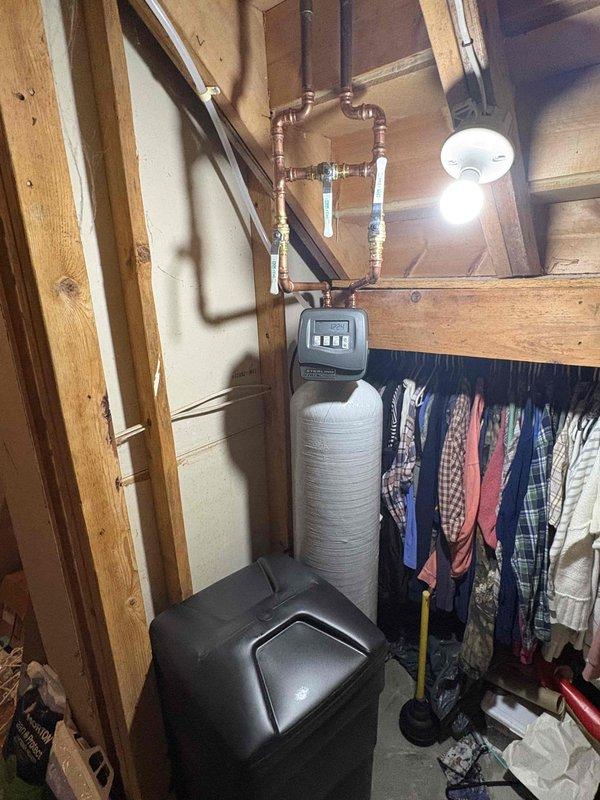 Completed installation of water softener system in basement utility area. Installed white cylindrical softener tank with insulated wrap, mounted gray digital control head with display panel, and connected copper supply lines from overhead joists to system with proper fittings and valves. Positioned brine tank receptacle on floor adjacent to softener unit and verified all plumbing connections for proper operation.