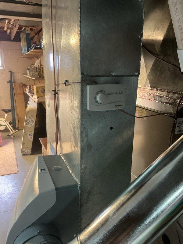 Completed installation of replacement humidifier unit with manual control in residential basement mechanical room. The new white humidifier unit was mounted adjacent to the existing green furnace and connected to the HVAC ductwork via flexible tubing. Installation included integration with the home's plumbing system, utilizing existing copper supply lines and brass fittings visible near the water heater assembly. All connections were secured, system was tested for proper operation, and the unit is now fully functional to provide whole-home humidity control.