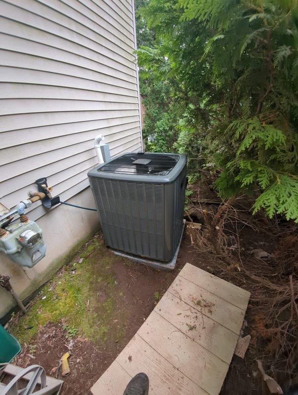 Completed full air conditioning system replacement at residential property. Removed existing outdoor condenser unit and installed new dark gray AC condenser on existing concrete pad along side of house near utility connections. Unit positioned in narrow access area between foundation and landscaping with proper clearance for airflow and service access. Verified all refrigerant line connections, electrical hookups, and drainage systems functioning properly. Confirmed indoor air handler integration with existing furnace and ductwork in basement mechanical room. All metallic ductwork connections inspected and secured. System tested for proper operation, temperature differential, and refrigerant charge levels. Unit operating within manufacturer specifications upon completion.