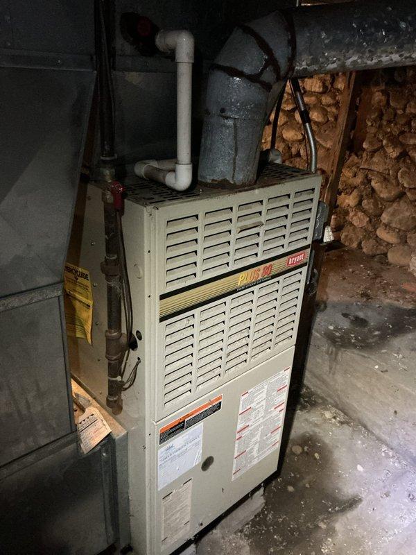 Responded to no cooling complaint on 15-year-old Bryant Plus 90 system. Upon arrival, confirmed blower motor failure preventing air circulation. Inspected furnace installation in basement utility area, noting unit positioned against stone foundation with standard PVC venting configuration. Diagnosed inoperative blower motor as primary cause of system failure. Documented equipment condition and installation setup including associated water heater and mechanical room layout for service records.