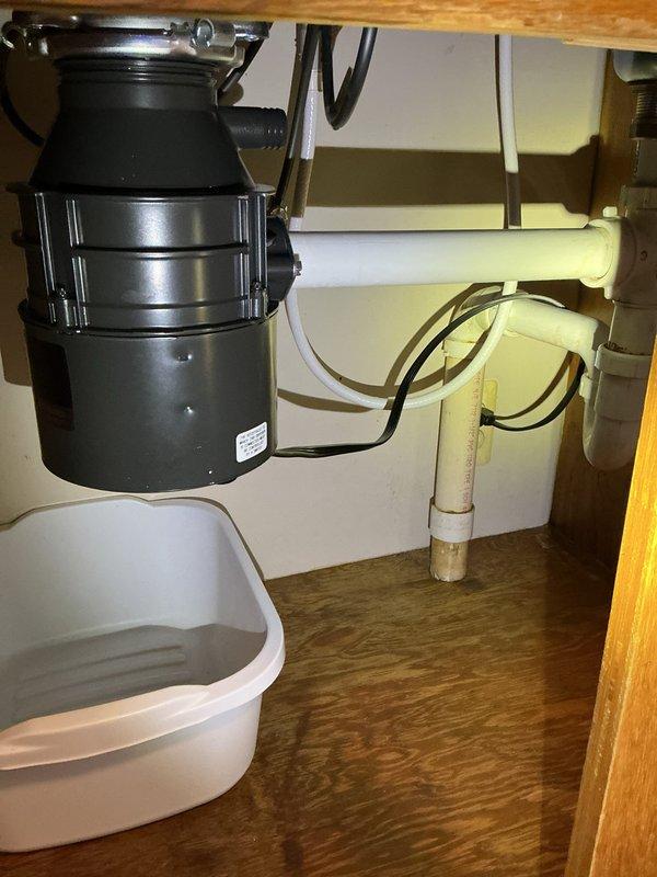 Responded to leaking garbage disposal under kitchen sink. Upon arrival, found active leak with homeowner using small pan to catch dripping water. Inspected disposal unit and surrounding plumbing connections under sink. Identified source of leak at disposal mounting assembly or drain connections. Documented existing conditions with photos showing black garbage disposal unit with metallic ribbed body, white PVC P-trap configuration, and cabinet interior setup. Assessed disposal unit condition, checked mounting flange, drain connections, and dishwasher inlet for proper seal and tightness. Resolved leak issue and verified no further water discharge from unit or connections. Tested disposal operation to ensure proper function after repair. Left work area clean and dry with leak eliminated.
