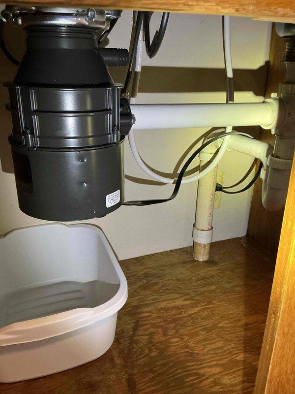 Responded to leaking garbage disposal under kitchen sink. Upon arrival, found active leak with homeowner using small pan to catch dripping water. Inspected disposal unit and surrounding plumbing connections under sink. Identified source of leak at disposal mounting assembly or drain connections. Documented existing conditions with photos showing black garbage disposal unit with metallic ribbed body, white PVC P-trap configuration, and cabinet interior setup. Assessed disposal unit condition, checked mounting flange, drain connections, and dishwasher inlet for proper seal and tightness. Resolved leak issue and verified no further water discharge from unit or connections. Tested disposal operation to ensure proper function after repair. Left work area clean and dry with leak eliminated.