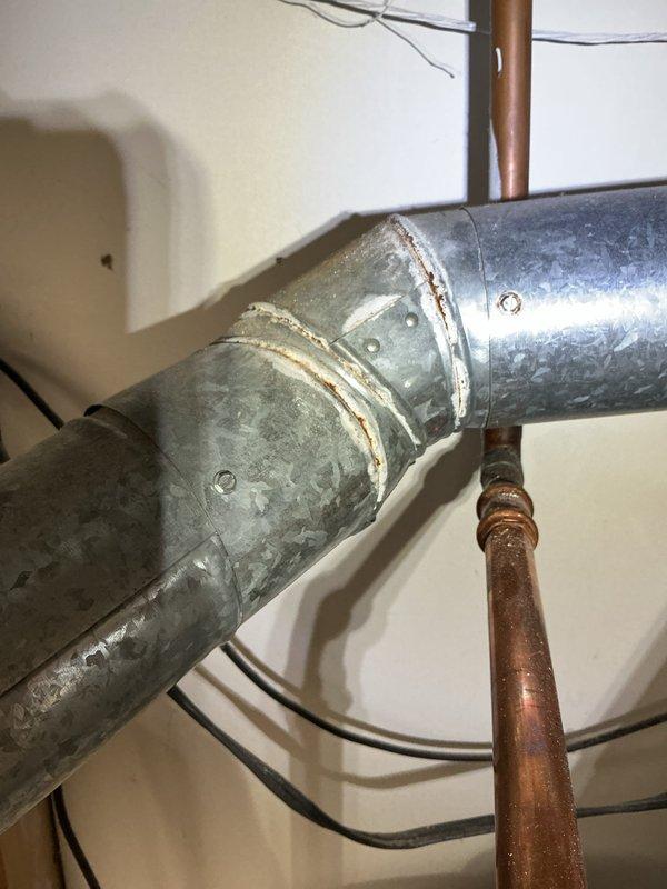 Responded to water leak service call where condensation drain line was actively leaking in the mechanical space. Upon inspection, identified compromised condensation drain line requiring attention to prevent continued water damage. Additionally observed that the galvanized flue pipe exhibited deteriorated seam joints showing signs of age, oxidation, and separation that required sealing work to ensure proper venting and prevent potential safety hazards. The flue pipe displayed significant weathering with visible surface corrosion and mottled patina throughout the ductwork sections. Documented existing conditions of HVAC system components including aged galvanized ductwork with riveted seams, copper refrigerant lines showing oxidation, and water heater installation with associated plumbing connections. System age could not be determined from available information on equipment. Photographed condensation drain line leak location and flue pipe seam deterioration for service records and futu