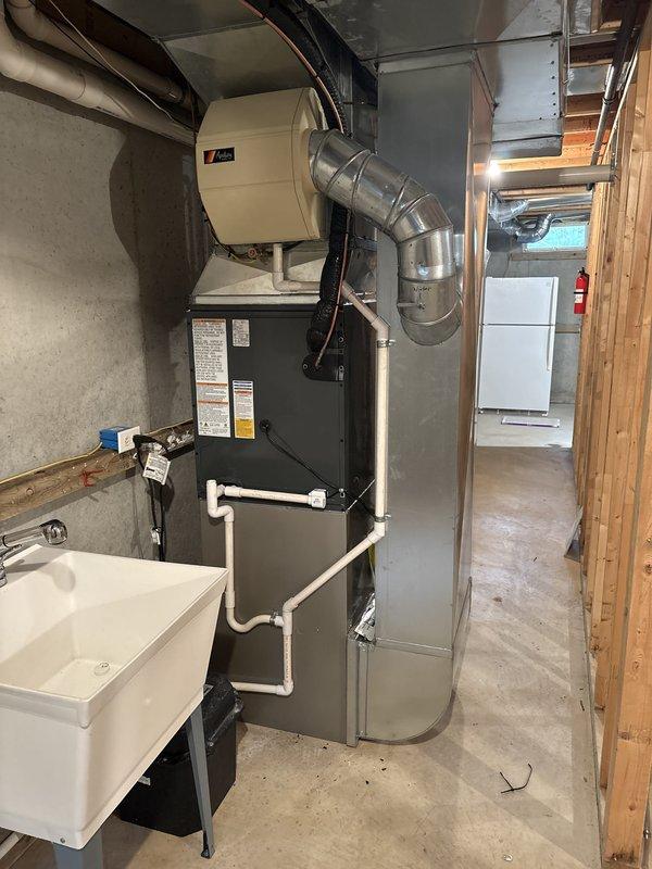 Completed full air conditioning system replacement in residential basement mechanical room. Installed new Carrier HVAC unit with integrated furnace, connected metallic ductwork with proper routing and elbow joints for optimal airflow distribution, and ensured all manufacturer labels and safety warnings were properly displayed. Verified all electrical connections, drainage systems via white PVC piping, and integration with existing humidifier unit mounted above the furnace. System tested and confirmed operational with proper airflow through ventilated panels and ductwork connections to adjacent air handler unit with filtration system.