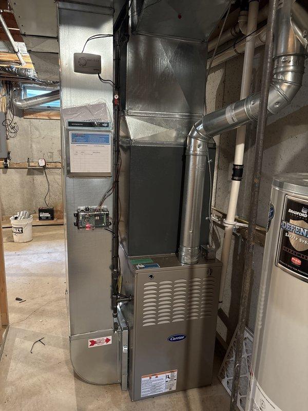 Completed full air conditioning system replacement in residential basement mechanical room. Installed new Carrier HVAC unit with integrated furnace, connected metallic ductwork with proper routing and elbow joints for optimal airflow distribution, and ensured all manufacturer labels and safety warnings were properly displayed. Verified all electrical connections, drainage systems via white PVC piping, and integration with existing humidifier unit mounted above the furnace. System tested and confirmed operational with proper airflow through ventilated panels and ductwork connections to adjacent air handler unit with filtration system.