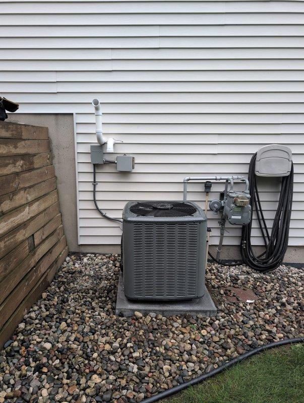 Completed full HVAC system replacement including outdoor condensing unit and indoor air handler/furnace. Removed existing equipment and installed new dark-colored condensing unit on existing concrete pad with proper refrigerant line sets, electrical disconnect, and PVC line hide. All refrigerant lines professionally routed up exterior wall with white line set covers for clean appearance. Indoor unit installed in basement utility area with new PVC venting system routed to exterior, secured with appropriate clamps and hangers. Connected all electrical components including disconnect box mounted to exterior wall per code requirements. System positioned on decorative river rock base with landscape edging for drainage and aesthetics. Completed all refrigerant connections, pressure tested system, evacuated lines, and charged to manufacturer specifications. Verified proper electrical connections, tested all safety controls, and confirmed system operation through complete heating and cooling c