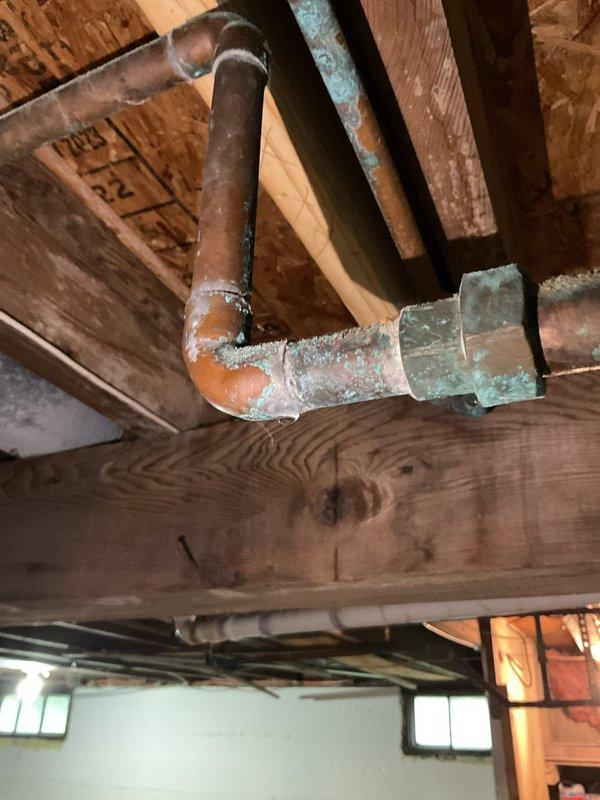 Responded to report of small leak above water heater in property being prepared for sale. Upon inspection, discovered corroded copper plumbing with significant verdigris oxidation on pipe joints and fittings in basement ceiling area. Documented existing conditions including aged copper piping system with green patina corrosion, exposed joists, and deteriorated plumbing infrastructure. Photographed corroded sections showing oxidation at elbow joints and connections, as well as overall utility space conditions including concrete block foundation walls and basement plumbing fixtures. Provided visual documentation of plumbing system age and condition for client's real estate transaction records.