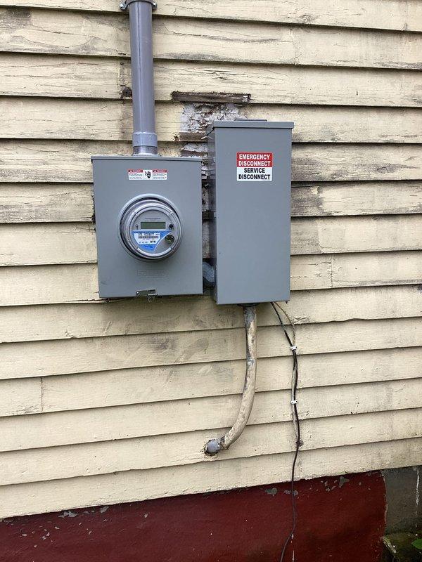 Responded to emergency service call for downed electrical mast. Upon arrival, found existing meter base and emergency disconnect panel mounted on exterior wall with gray PVC conduit extending upward. Inspected weatherhead and service entrance conductors for damage from mast failure. Documented existing water damage and staining on siding between meter and disconnect panel, indicating previous moisture infiltration. Evaluated vertical conduit mounting and bracket integrity along building exterior. Photographed current installation showing digital meter, emergency disconnect box with proper labeling, and conduit routing. Assessed weathering patterns and dark streaking on vinyl siding suggesting long-term water runoff from roofline. Examined soffit area where service entrance cables enter structure, noting cable condition and support. Verified proper grounding and bonding connections at meter base. Restored electrical service safely and confirmed system operational.
