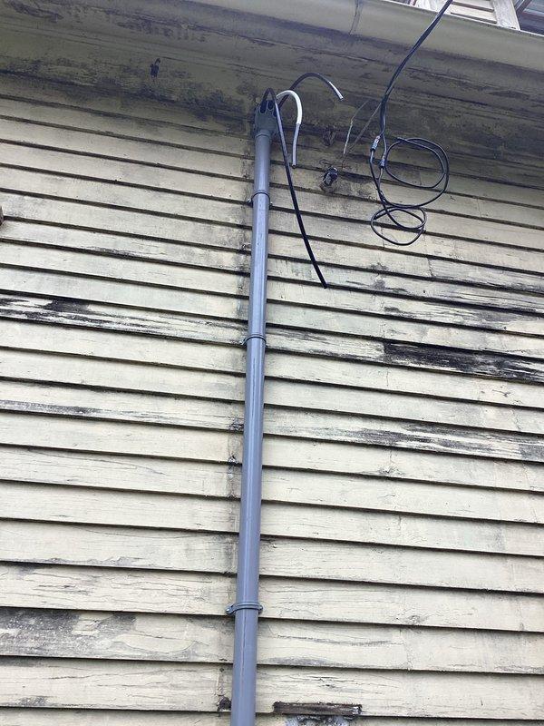 Responded to emergency service call for downed electrical mast. Upon arrival, found existing meter base and emergency disconnect panel mounted on exterior wall with gray PVC conduit extending upward. Inspected weatherhead and service entrance conductors for damage from mast failure. Documented existing water damage and staining on siding between meter and disconnect panel, indicating previous moisture infiltration. Evaluated vertical conduit mounting and bracket integrity along building exterior. Photographed current installation showing digital meter, emergency disconnect box with proper labeling, and conduit routing. Assessed weathering patterns and dark streaking on vinyl siding suggesting long-term water runoff from roofline. Examined soffit area where service entrance cables enter structure, noting cable condition and support. Verified proper grounding and bonding connections at meter base. Restored electrical service safely and confirmed system operational.