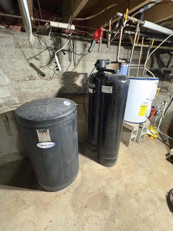 Responded to water heater odor complaint potentially related to anode rod issues. Upon arrival, inspected residential water heater installation located in basement utility area alongside water treatment equipment. Performed visual assessment of water heater condition, noting standard copper piping connections and brass fittings at top of unit. Examined anode rod as primary suspect for sulfur or rotten egg odor commonly associated with bacterial reaction in tank. Documented equipment configuration including proximity to water softener system and filtration equipment, which can contribute to anode rod deterioration and odor problems. Photographed installation for service records showing overall mechanical room layout and close-up views of water heater connections and top access point. Provided customer with findings and recommendations regarding anode rod replacement or alternative solutions to resolve odor issue.
