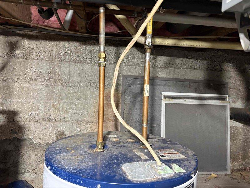 Responded to water heater odor complaint potentially related to anode rod issues. Upon arrival, inspected residential water heater installation located in basement utility area alongside water treatment equipment. Performed visual assessment of water heater condition, noting standard copper piping connections and brass fittings at top of unit. Examined anode rod as primary suspect for sulfur or rotten egg odor commonly associated with bacterial reaction in tank. Documented equipment configuration including proximity to water softener system and filtration equipment, which can contribute to anode rod deterioration and odor problems. Photographed installation for service records showing overall mechanical room layout and close-up views of water heater connections and top access point. Provided customer with findings and recommendations regarding anode rod replacement or alternative solutions to resolve odor issue.