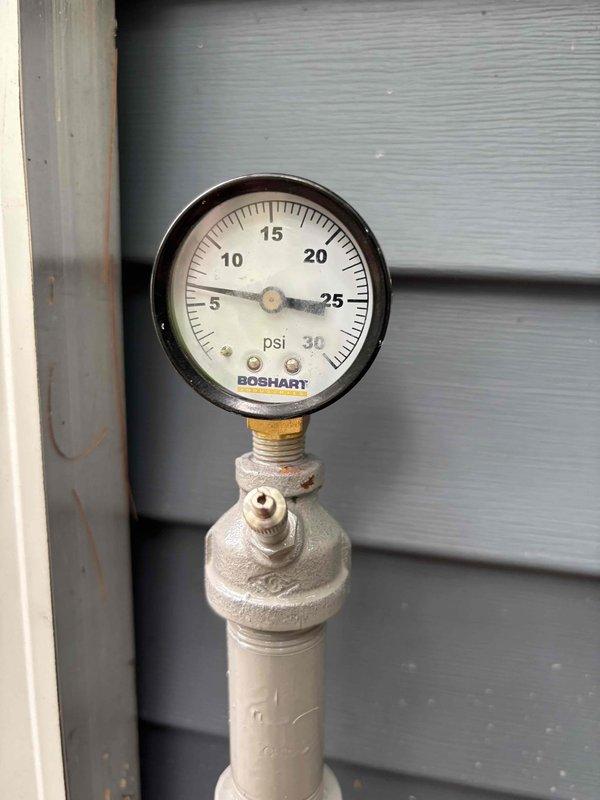 Removed Boshart brand pressure gauge (0-30 psi range) from exterior gas line pipe connection. Gauge was mounted on painted pipe against building exterior siding via brass threaded fitting. After removal, properly capped the pipe connection point to seal the system. PVC utility pipe configuration includes multiple couplings and fittings with valve assembly at top, positioned at ground level near building foundation.