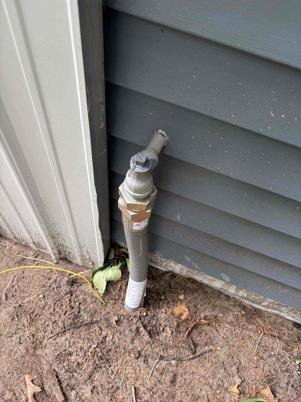 Removed Boshart brand pressure gauge (0-30 psi range) from exterior gas line pipe connection. Gauge was mounted on painted pipe against building exterior siding via brass threaded fitting. After removal, properly capped the pipe connection point to seal the system. PVC utility pipe configuration includes multiple couplings and fittings with valve assembly at top, positioned at ground level near building foundation.