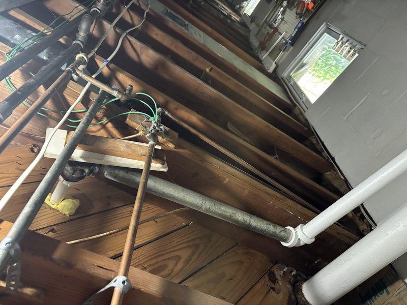 Responded to service call for leaking water heater in single-family residence. Upon arrival, inspected 7-year-old standard tank water heater and associated plumbing connections in attic space. Located active leak source and assessed surrounding copper supply lines, PVC drainage piping, and electrical connections for potential contributing factors. Documented existing plumbing infrastructure including shut-off valves, supply lines, and drain configurations. Photographed attic installation showing water heater placement, pipe routing through wooden beam structure, and proximity to electrical junction boxes. Also documented bathroom plumbing connections and emergency water collection measures in place. Completed visual inspection of all accessible components to determine extent of leak and recommend appropriate repair or replacement options to homeowner.