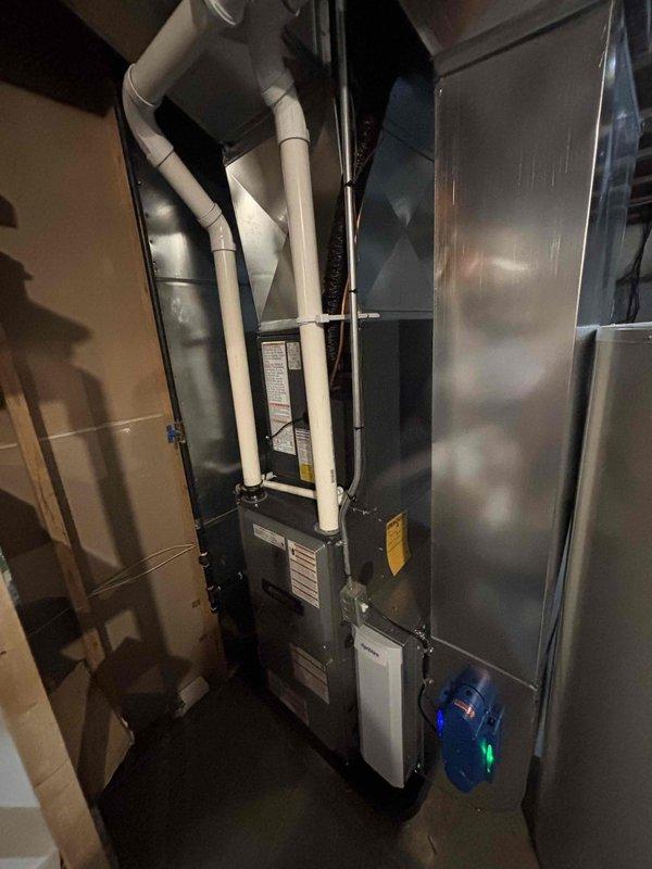 Completed full installation of residential HVAC system including new furnace and air conditioning unit in basement mechanical room. Installed dark-colored high-efficiency furnace with proper white PVC venting routed vertically through structure. Mounted gray metal air handler unit adjacent to furnace with metallic supply ductwork connections. Installed system monitoring and control device with operational indicators at base of equipment. Secured all PVC piping with appropriate brackets and straps to ensure code-compliant installation. Verified all electrical connections, tested system operation, and confirmed proper airflow through ductwork. System powered on successfully with all safety controls and monitoring devices functioning as designed.