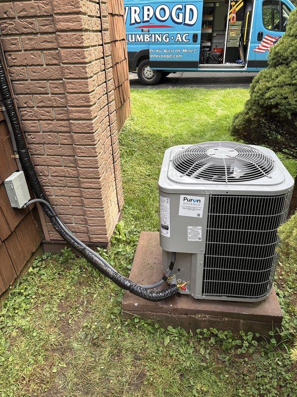 Completed full air conditioning system replacement at residential property. Removed existing outdoor condenser unit and installed new Puron-compatible AC condenser on concrete pad adjacent to brick exterior, connecting refrigerant lines via black flexible conduit from house to unit. Installed new indoor air handler in utility room with proper ductwork connections, including silver metallic supply plenum and trunk lines. Integrated system with existing furnace infrastructure, ensuring proper electrical connections through dedicated disconnect boxes and control panels mounted to wall studs. Verified all refrigerant line connections, electrical wiring through conduit, and gas line shut-off valves for safe operation. System tested and confirmed operational with proper airflow and cooling performance.