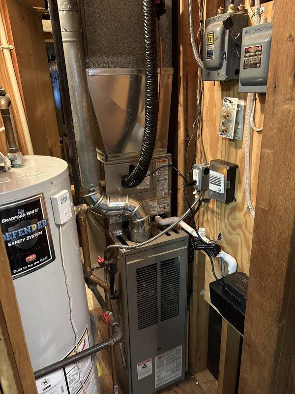 Completed full air conditioning system replacement at residential property. Removed existing outdoor condenser unit and installed new Puron-compatible AC condenser on concrete pad adjacent to brick exterior, connecting refrigerant lines via black flexible conduit from house to unit. Installed new indoor air handler in utility room with proper ductwork connections, including silver metallic supply plenum and trunk lines. Integrated system with existing furnace infrastructure, ensuring proper electrical connections through dedicated disconnect boxes and control panels mounted to wall studs. Verified all refrigerant line connections, electrical wiring through conduit, and gas line shut-off valves for safe operation. System tested and confirmed operational with proper airflow and cooling performance.