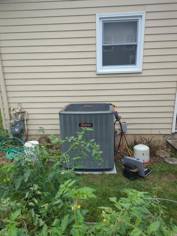 Completed full air conditioning system replacement at residential property. Removed existing outdoor condensing unit and installed new Amana outdoor unit adjacent to building on mulch bed near gas meter. Performed refrigerant line connections using brass fittings and copper piping with proper insulation, verified all connections secure and leak-free. Connected electrical conduit and drain line, applied service identification labels showing system specifications and refrigerant charge information. System tested and operational upon completion.