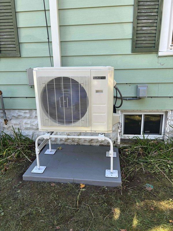 Completed installation of DURASTAR ductless mini-split system including outdoor condensing unit mounted on platform bracket system with white support legs anchored to base pad, and indoor wall-mounted air handler. Installed refrigerant line set running vertically along exterior siding from outdoor unit to indoor unit, secured electrical conduit and wiring connections, and mounted electrical junction box on exterior wall. System tested and operational upon completion.