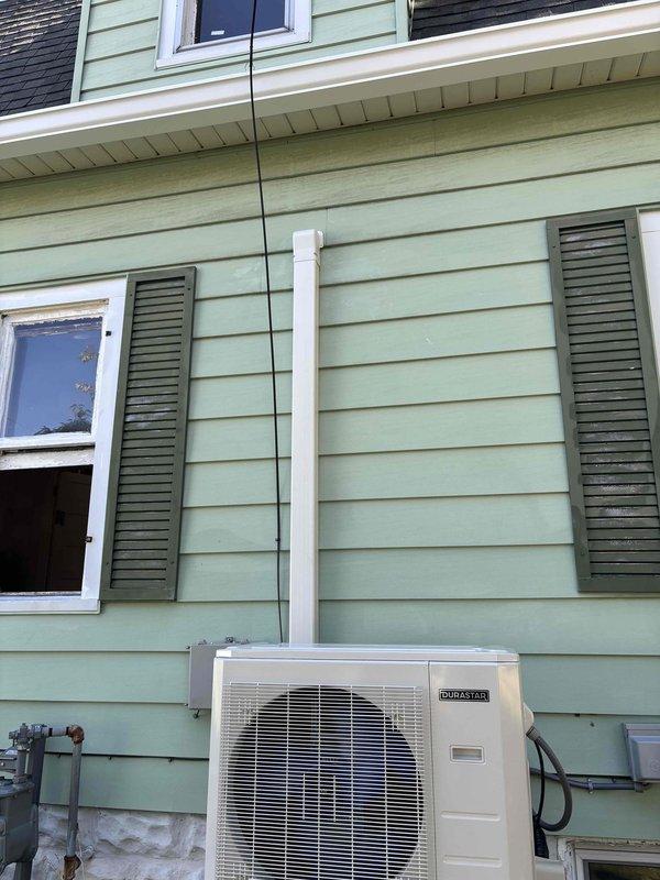 Completed installation of DURASTAR ductless mini-split system including outdoor condensing unit mounted on platform bracket system with white support legs anchored to base pad, and indoor wall-mounted air handler. Installed refrigerant line set running vertically along exterior siding from outdoor unit to indoor unit, secured electrical conduit and wiring connections, and mounted electrical junction box on exterior wall. System tested and operational upon completion.