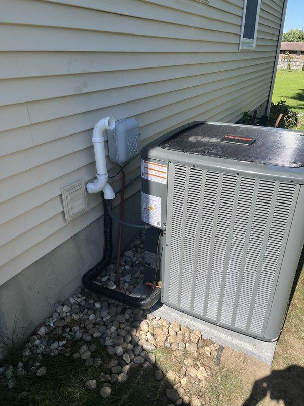 Completed full residential HVAC system installation including new furnace and air conditioning unit. Installed gray metal furnace with evaporator coil in basement utility area on black drain pan with proper condensate drainage via white PVC piping and venting lines. Mounted outdoor AC condenser unit on concrete pad adjacent to building exterior with white PVC refrigerant line connections through wall penetration. Connected black flexible ductwork and refrigerant lines between indoor and outdoor components. Installed wall-mounted thermostat control unit for system operation. Verified all electrical connections, tested system startup and operation, and confirmed proper airflow through metal ductwork. System is fully operational and ready for use.