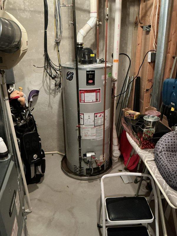 Responded to no hot water call and found 6-year-old standard tank water heater leaking from top ring with water running down the side of unit. Leak was contained in drain pan with no water damage to basement floor. Documented equipment condition with photos showing water heater installation, plumbing connections, and overhead piping configuration including PVC discharge lines and copper supply lines.
