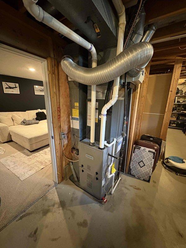 Completed full air conditioning system replacement at residential property. Removed existing outdoor condenser unit and installed new gray-green condensing unit against exterior wall with proper refrigerant line connections and electrical hookup. Unit positioned on stable pad with appropriate clearances from landscaping and foundation. Connected system to existing basement furnace and ductwork infrastructure, ensuring proper integration with forced air distribution system. Verified all refrigerant lines secured, electrical connections completed to code, and condensate drainage functioning properly. System tested and confirmed operational with proper airflow and cooling capacity.