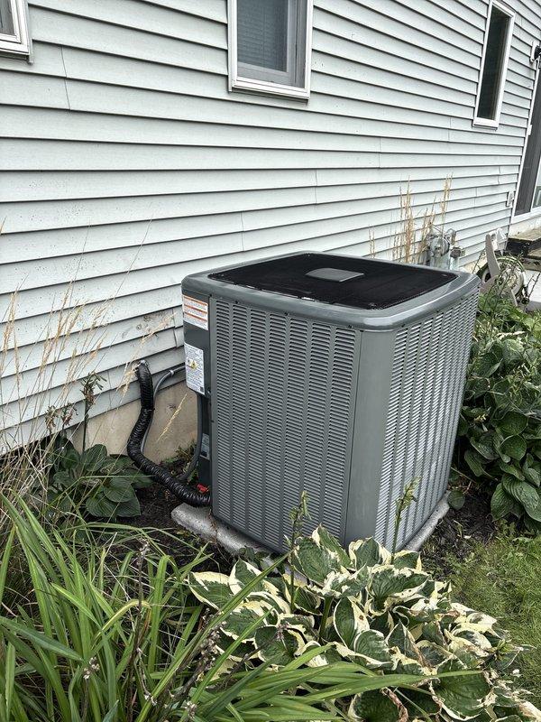 Completed full air conditioning system replacement at residential property. Removed existing outdoor condenser unit and installed new gray-green condensing unit against exterior wall with proper refrigerant line connections and electrical hookup. Unit positioned on stable pad with appropriate clearances from landscaping and foundation. Connected system to existing basement furnace and ductwork infrastructure, ensuring proper integration with forced air distribution system. Verified all refrigerant lines secured, electrical connections completed to code, and condensate drainage functioning properly. System tested and confirmed operational with proper airflow and cooling capacity.