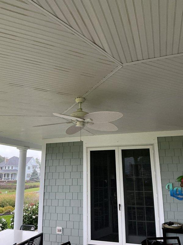 Responded to service call for non-functioning outdoor ceiling fan located on covered patio area with beadboard ceiling and green shingle siding. Removed existing failed ceiling fan unit from mounting bracket. Installed new outdoor-rated replacement ceiling fan with woven wicker-style blades and sage-colored motor housing, securing to existing electrical box and downrod. Verified proper electrical connections, tested fan operation at all speeds, and confirmed pull chain functionality. Customer was not present during service visit; accessed property using provided gate and garage codes.