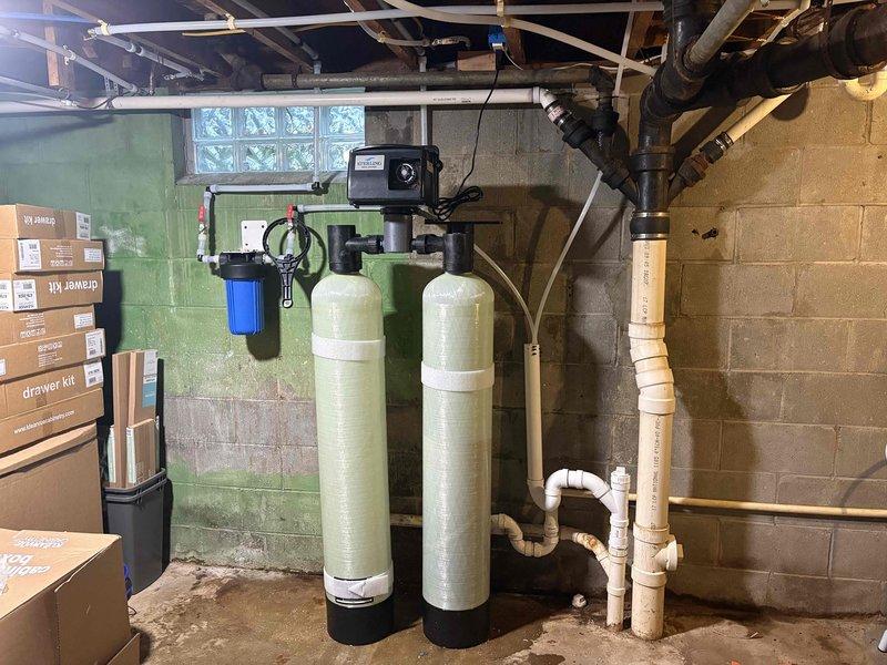 Completed installation of iron curtain water treatment system in basement utility area. System includes two tall cylindrical filtration tanks with black control unit, blue filter housing, and associated valves and piping mounted on concrete block wall. Connected copper and PVC plumbing lines to integrate system with existing water supply infrastructure.