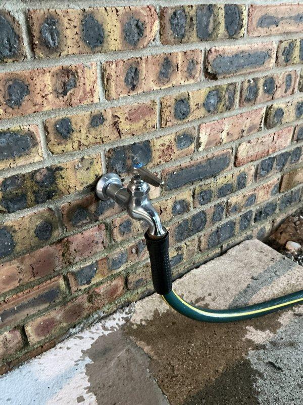 Inspected two outdoor faucets showing active leaks and provided repair/replacement quote as requested. The first faucet, a chrome cross-handle spigot mounted on brick exterior, exhibited leakage evidenced by white mineral deposits and water staining on concrete below. The second location featured a backflow prevention assembly with multiple valves on gray siding, also connected to garden hose with visible moisture concerns on surrounding pavers.