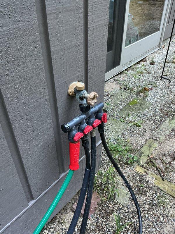 Inspected two outdoor faucets showing active leaks and provided repair/replacement quote as requested. The first faucet, a chrome cross-handle spigot mounted on brick exterior, exhibited leakage evidenced by white mineral deposits and water staining on concrete below. The second location featured a backflow prevention assembly with multiple valves on gray siding, also connected to garden hose with visible moisture concerns on surrounding pavers.