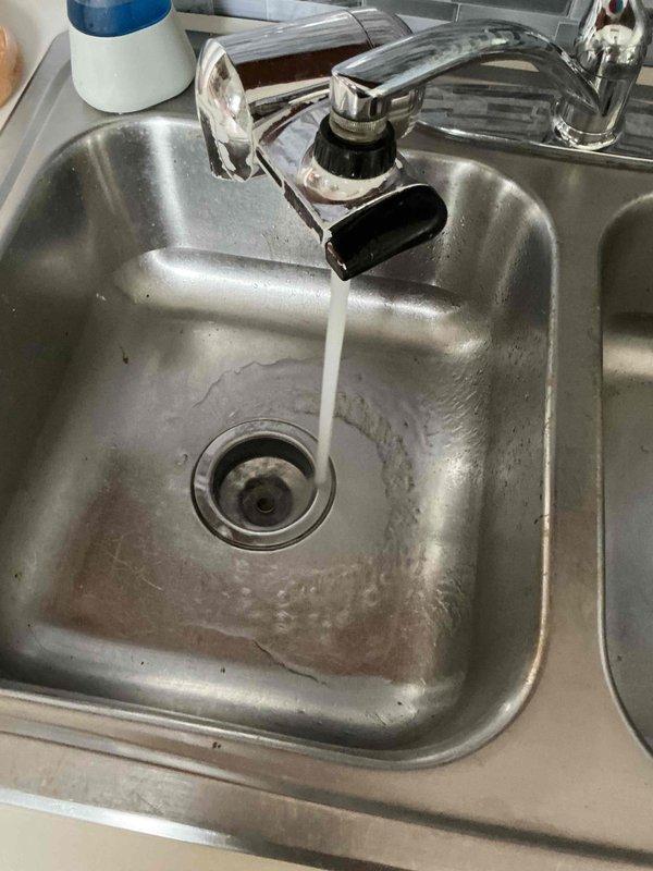 Responded to overflowing kitchen sink issue in residential kitchen. Upon inspection, found double-basin stainless steel sink with drainage concerns and mineral deposit buildup around drain assembly. Accessed plumbing system beneath sink through crawlspace access point, where aging pipes and connections showed signs of deterioration. Cleared blockage, restored proper drainage function, and documented pre-existing conditions including water staining and worn plumbing components with metallic tape repairs.