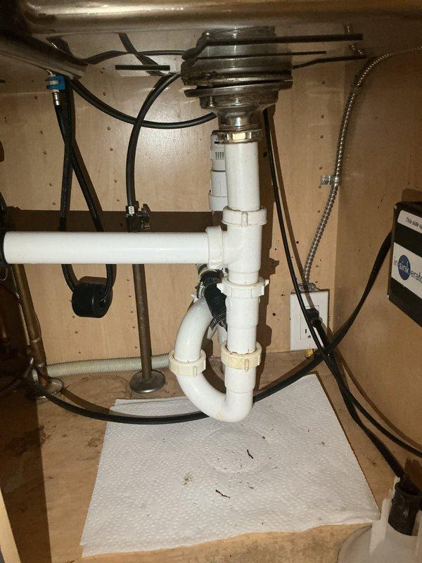 Responded to kitchen sink leak complaint. Upon inspection, identified leak originating from compromised connections in the PVC drain assembly beneath double-basin sink. The drainage system included a garbage disposal unit connected to white PVC P-trap configuration with multiple joints and elbow fittings. Inspected all visible plumbing components including disposal unit, drain connections, supply lines, and trap assembly. Tightened loose slip-joint connections at P-trap and verified proper sealing of disposal discharge connection to drain system. Tested all connections under running water to confirm leak was eliminated. Left area clean and dry with no active water discharge detected at completion.