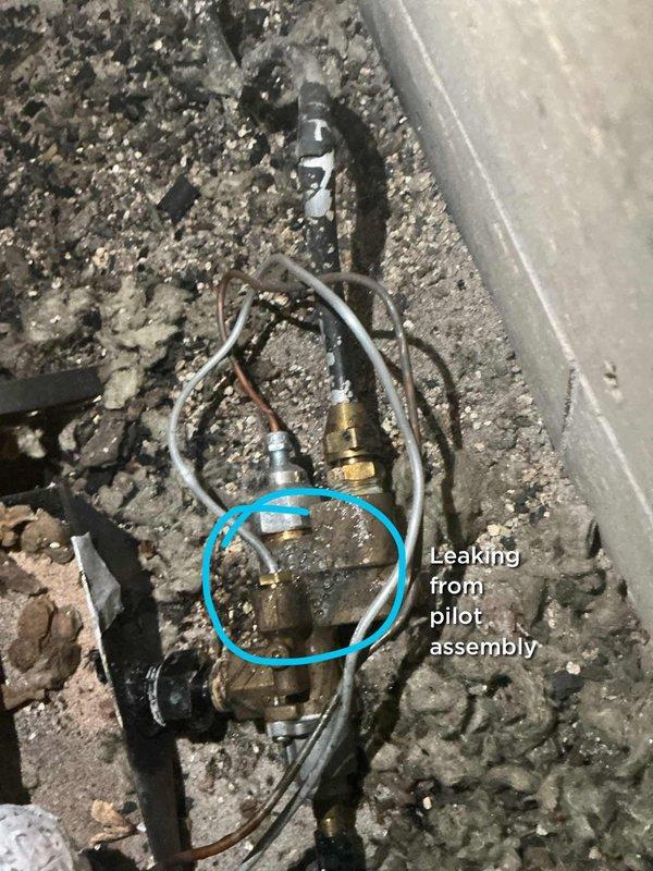 Responded to reported hissing sound from gas fireplace area in 1980s-era condo unit. Upon inspection, identified active gas leak at pilot assembly of original fireplace installation. Leak was isolated to brass fitting connection on pilot assembly mechanism. Heavy dust and debris accumulation noted throughout equipment compartment, consistent with 40+ years of service without maintenance. Documented leak location with photographic evidence showing compromised pilot assembly components. Advised on necessary repairs to pilot assembly to eliminate gas leak hazard and restore safe fireplace operation.