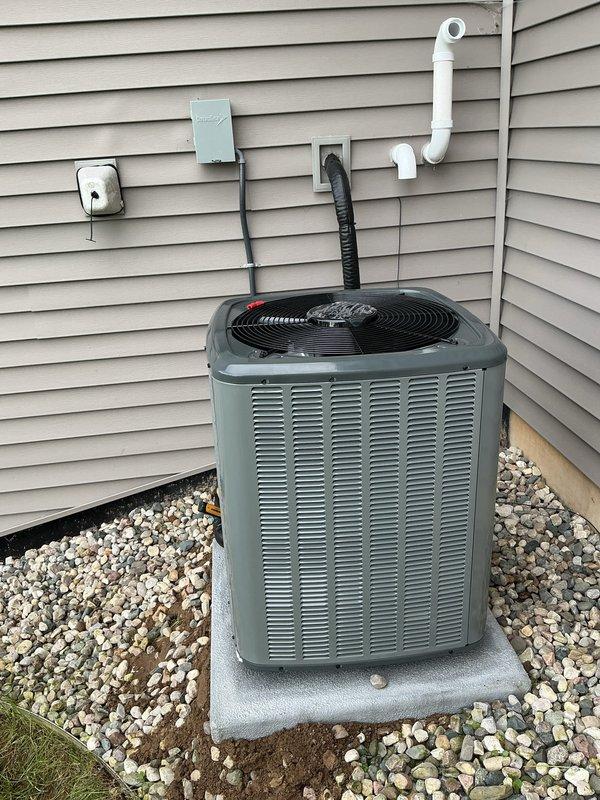 Completed full air conditioning system replacement at residential property. Removed existing outdoor condensing unit and installed new gray-green AC unit on concrete pad with decorative rock surround. Mounted electrical disconnect panel and outdoor outlet cover on exterior wall, ran black flexible conduit from wall to unit, and installed white PVC refrigerant line set with proper insulation. Inside mechanical room, installed new Lennox high-efficiency furnace with white PVC exhaust venting routed through ceiling. Connected furnace to existing ductwork using black flexible duct connections. Verified proper clearances around furnace and adjacent water heater. Completed all electrical connections, tested system operation, and confirmed proper airflow and refrigerant charge levels.