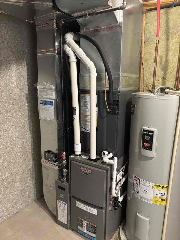 Completed full air conditioning system replacement at residential property. Removed existing outdoor condensing unit and installed new gray-green AC unit on concrete pad with decorative rock surround. Mounted electrical disconnect panel and outdoor outlet cover on exterior wall, ran black flexible conduit from wall to unit, and installed white PVC refrigerant line set with proper insulation. Inside mechanical room, installed new Lennox high-efficiency furnace with white PVC exhaust venting routed through ceiling. Connected furnace to existing ductwork using black flexible duct connections. Verified proper clearances around furnace and adjacent water heater. Completed all electrical connections, tested system operation, and confirmed proper airflow and refrigerant charge levels.