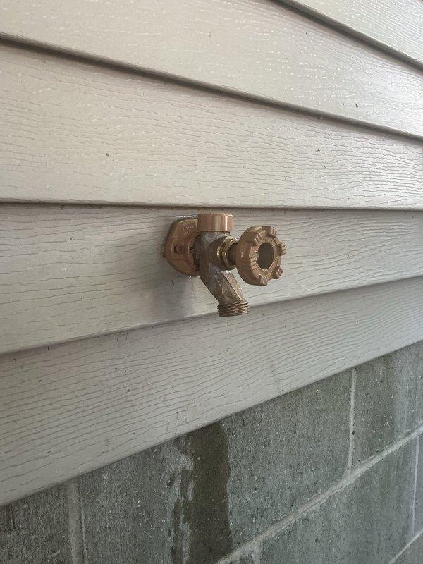Responded to service call for leaking outdoor hose bib. Upon inspection, found standard brass spigot with wheel-style handle valve mounted on exterior wall above foundation level showing signs of weathering, corrosion, and mineral deposits. Diagnosed leak source, performed necessary repairs to stop water discharge, and tested fixture to confirm proper operation. Documented completed work with photographs showing spigot assembly, wall mounting, and surrounding installation area.