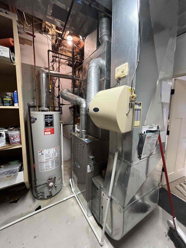 Completed full furnace installation in basement utility room. Installed new gray metal furnace unit with connected ductwork, integrated beige humidifier system with control panel, and routed all necessary ventilation ducts vertically to ceiling connections. All electrical connections completed, system tested and verified operational alongside existing water heater.