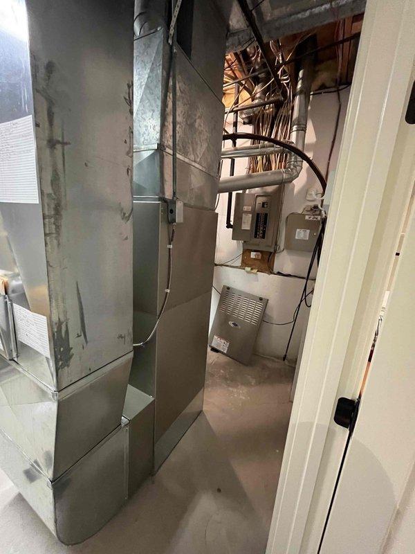 Completed full furnace installation in basement utility room. Installed new gray metal furnace unit with connected ductwork, integrated beige humidifier system with control panel, and routed all necessary ventilation ducts vertically to ceiling connections. All electrical connections completed, system tested and verified operational alongside existing water heater.