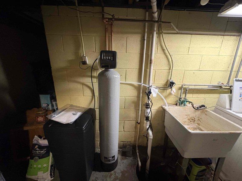 Completed installation and setup of water softener system in basement utility area. Connected copper supply lines to filtration unit with black digital control head, verified proper operation of all plumbing connections, and stocked salt supply for system maintenance.