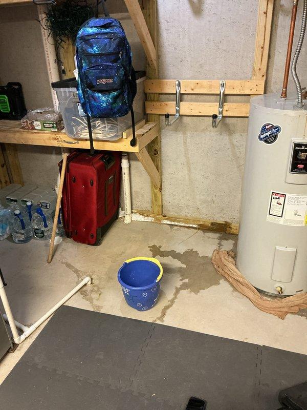 Responded to service call for reported water heater leak in basement utility area. Upon arrival, found active moisture and water staining on concrete floor surrounding white residential water heater unit. Inspected water heater connections, tank exterior, temperature and pressure relief valve, and drain valve for source of leak. Documented existing conditions with photographs showing water damage patterns on floor and temporary cloth barrier placed around base of unit. Assessed age and overall condition of water heater, noting manufacturer specifications and existing plumbing configuration with copper supply lines and flexible connector at top of tank.