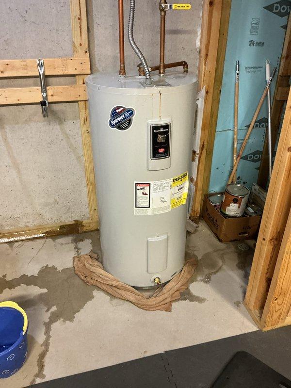 Responded to service call for reported water heater leak in basement utility area. Upon arrival, found active moisture and water staining on concrete floor surrounding white residential water heater unit. Inspected water heater connections, tank exterior, temperature and pressure relief valve, and drain valve for source of leak. Documented existing conditions with photographs showing water damage patterns on floor and temporary cloth barrier placed around base of unit. Assessed age and overall condition of water heater, noting manufacturer specifications and existing plumbing configuration with copper supply lines and flexible connector at top of tank.